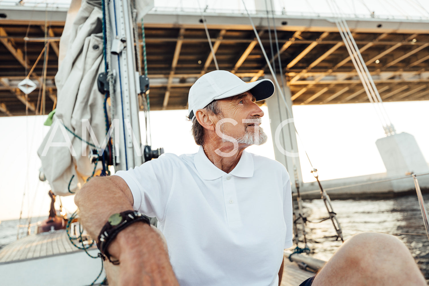 Relaxed mature man sitting on his yacht. Senior male in cap looking away while floating under a highway on a sailboat.