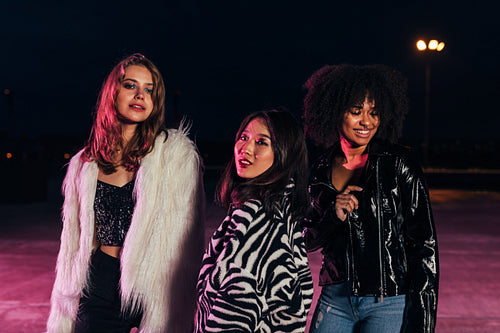 Fashionable girls standing together on roof at night under neon lights. Multi-ethnic group of women posing outdoors and looking at camera.