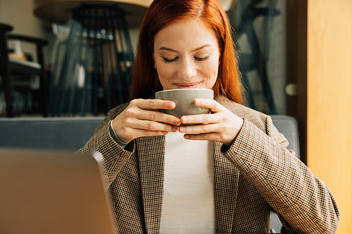 Close-up of a young businesswoman holding a cup and smiling. Female with ginger hair enjoying her coffee in the morning in a cafe.