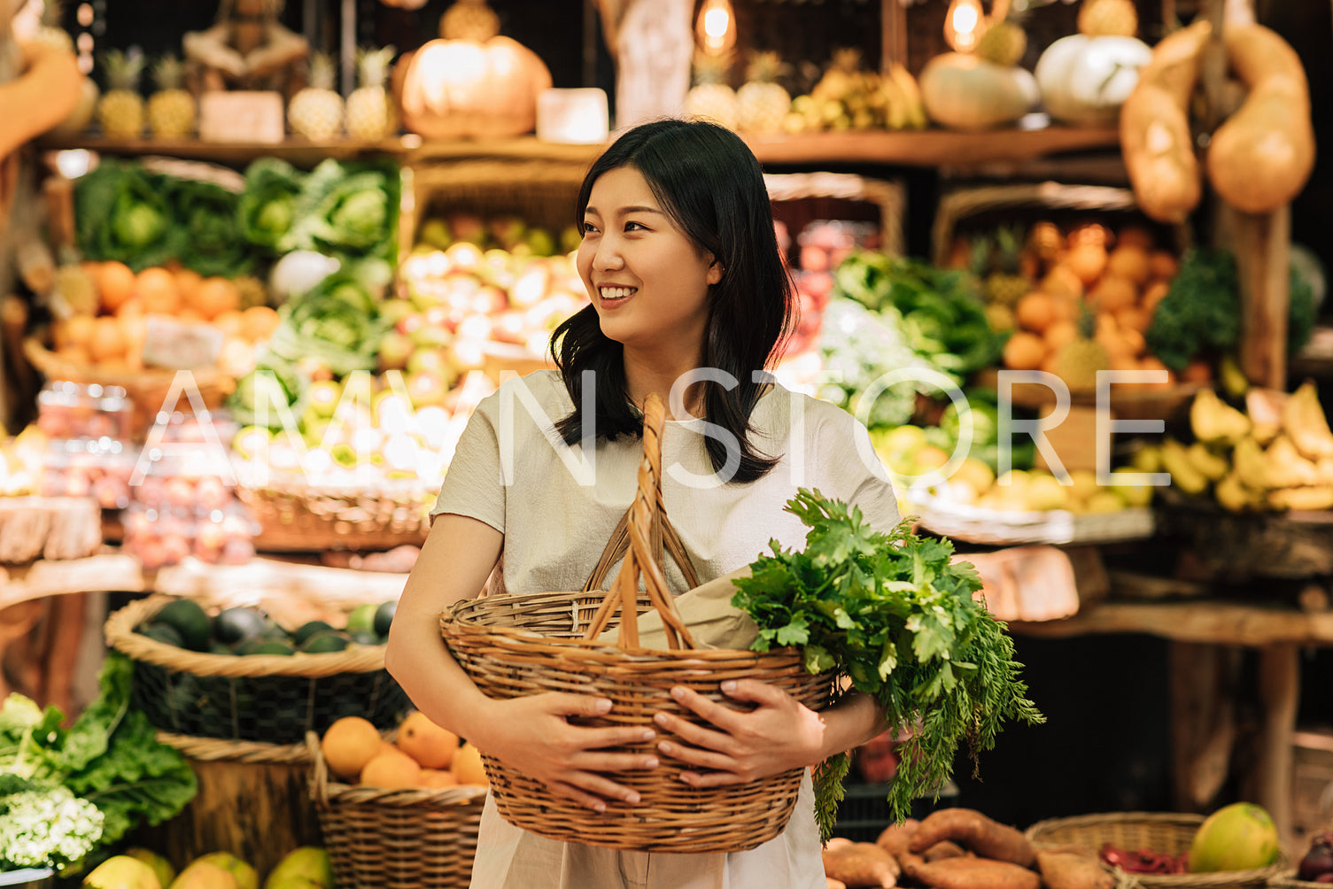 Smiling Asian woman holding wicker shopping basket with vegetables while standing at the farmers market