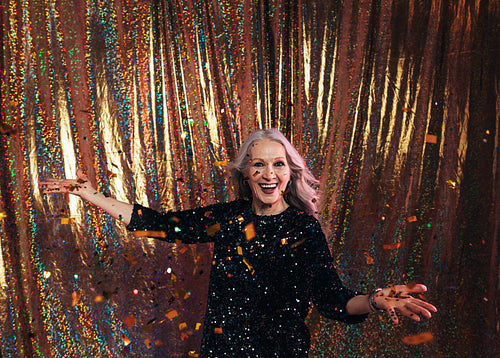 Happy senior woman in black dress standing under confetti agains