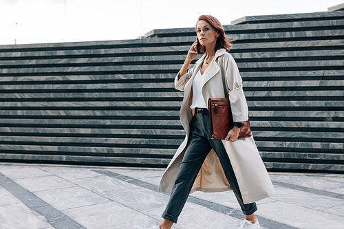 Stylish woman in a coat walking outdoors talking on a mobile phone. Businesswoman with ginger hair and a leather folder walking on the street.