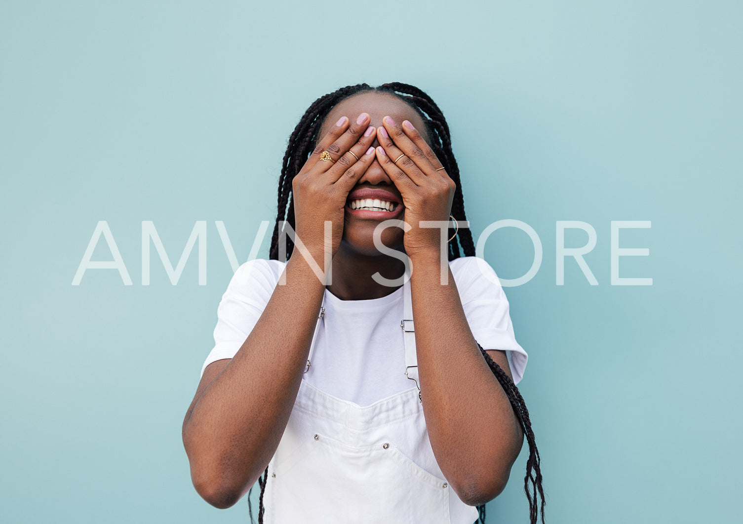 Smiling female with braids hiding her face with palms while standing at a blue wall outdoors