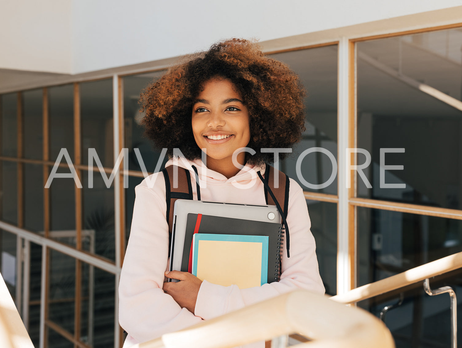 Smiling teenage girl with books and laptop standing on stairs in college and looking away