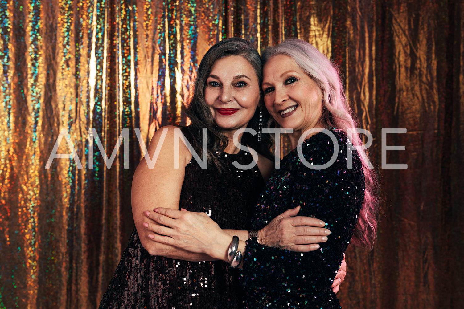 Two smiling senior girlfriends wearing evening dresses hugging each other while standing at glitter background