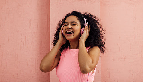 Laughing woman listening to music with pink headphones with closed eyes. Young happy female in pink casuals enjoying music.