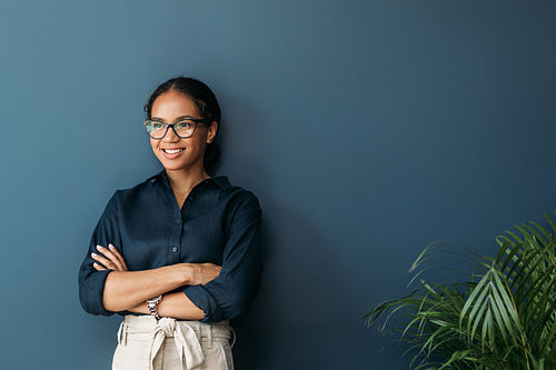 Confident businesswoman with crossed arms leaning on blue wall. Smiling entrepreneur in eyeglasses looking away.