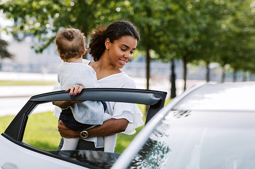 Woman opening a car door and putting her little daughter into it. Young female holding little girl on hands near a car.