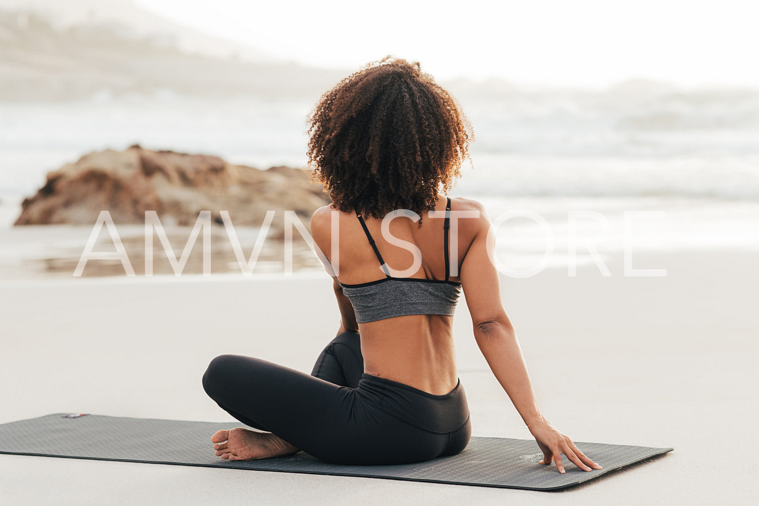 Rear view of slim woman sitting on a mat at beach and looking at ocean while exercising