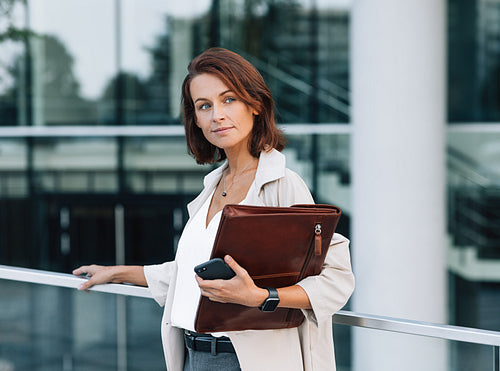 Confident middle-aged woman in stylish clothes standing outdoors. Female with ginger hair holding a leather folder while standing against an office building.