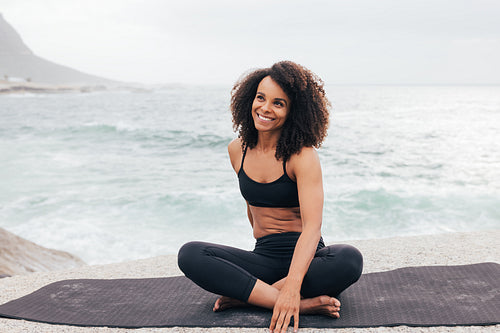 Portrait of young smiling female in sportswear sitting on yoga mat by ocean