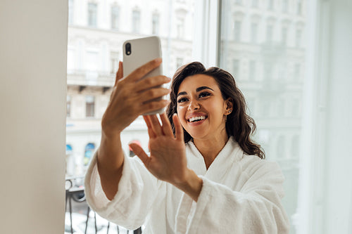 Happy beautiful woman taking a selfie on a balcony in her hotel room. Smiling female in bathrobe holding smartphone and making photographs on frontal camera.