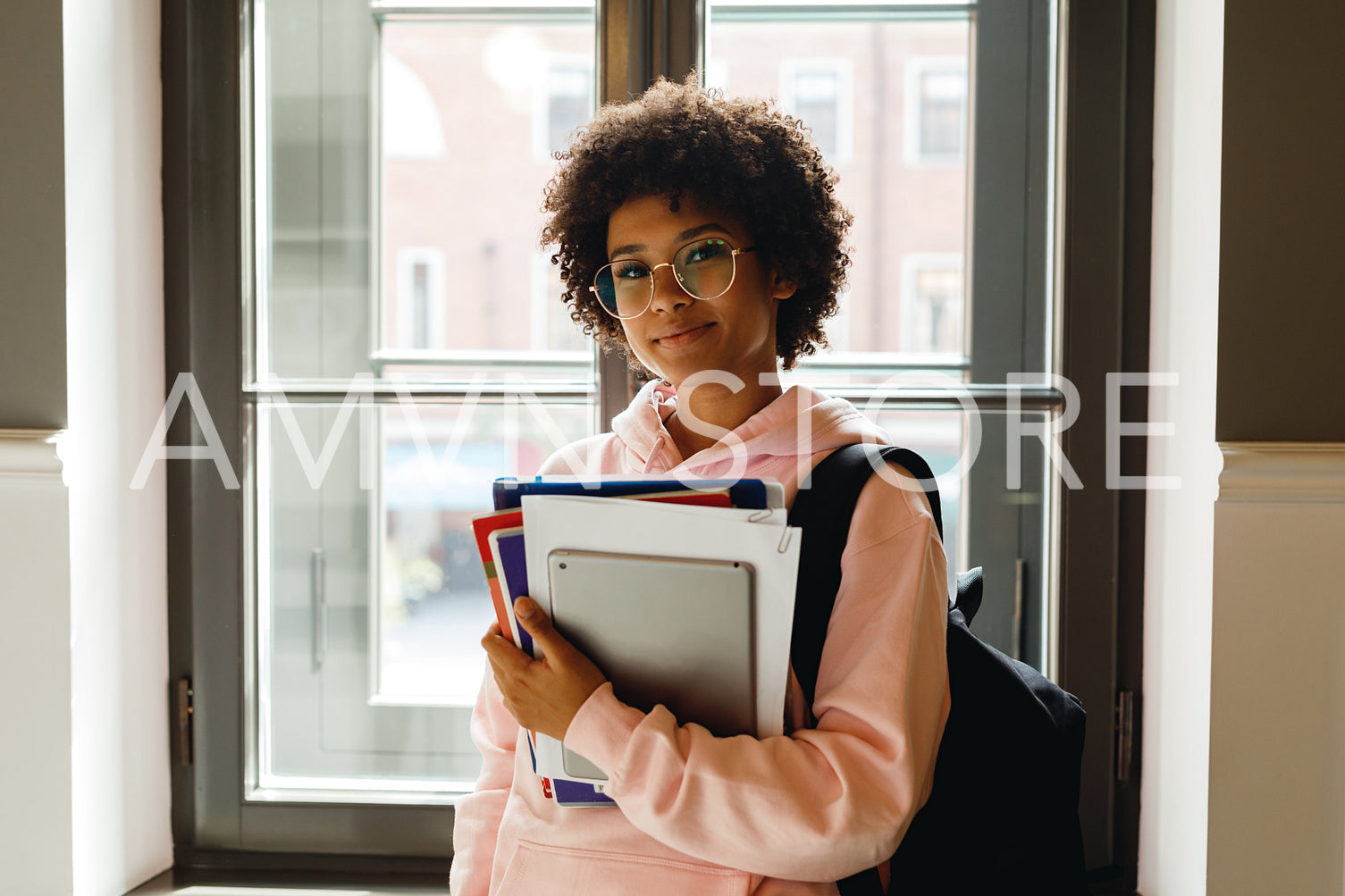 Beautiful young woman with books and digital tablet standing at window in campus	