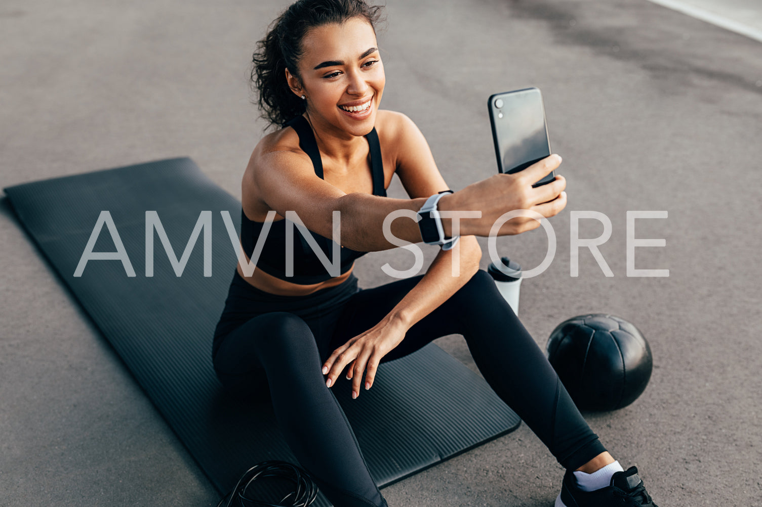 Female athlete sitting on a mat outdoors and taking selfie for social media. Smiling woman taking a break during training in the evening.	