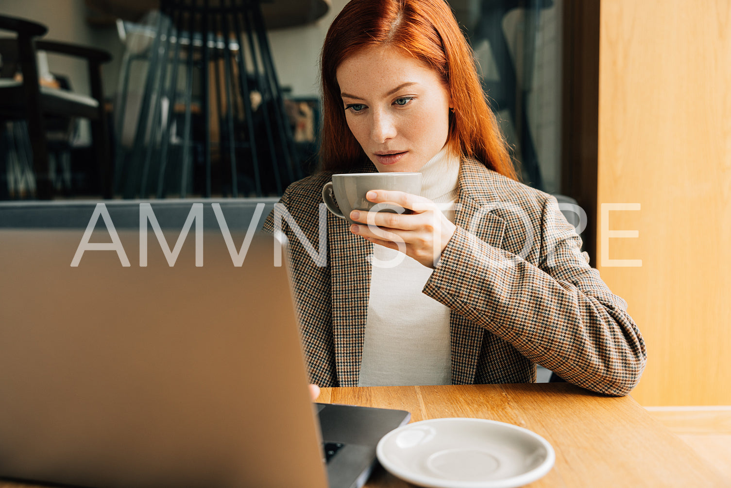 Young woman with ginger hair holding a cup of coffee and working on a laptop in cafe