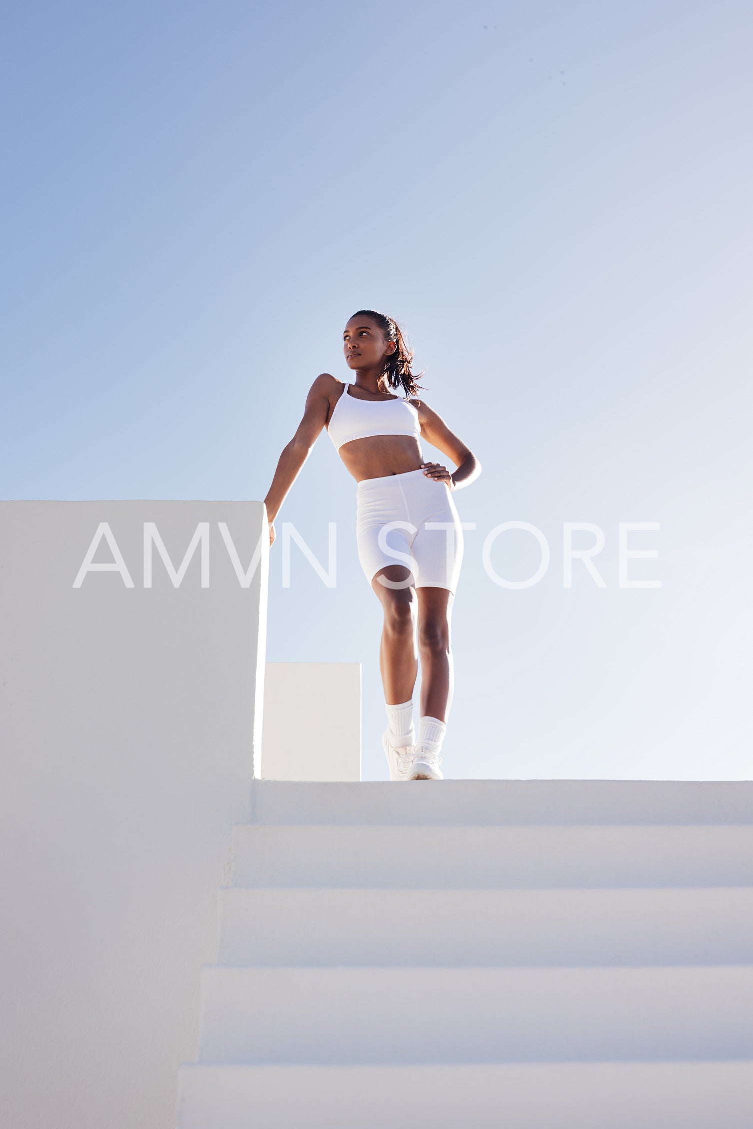 Full length of a young slim female posing while standing on the top of stairs. Woman in white fitness attire standing outdoors against the skies.