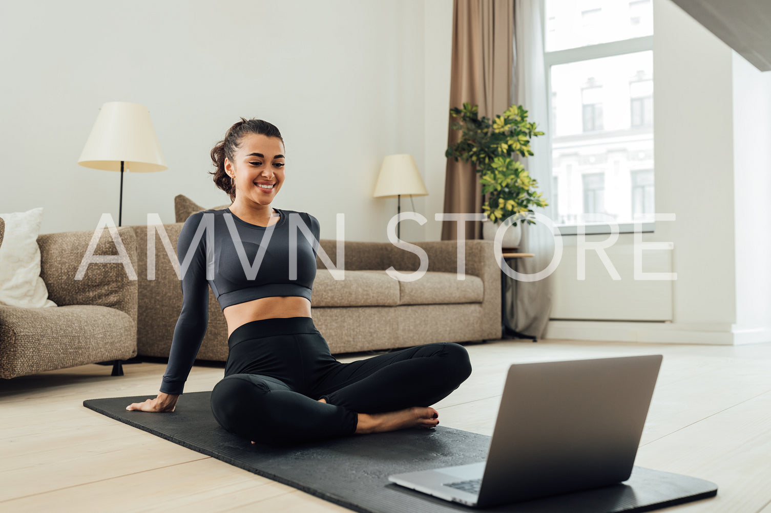 Smiling woman in sportswear sitting on a mat in the living room looking at laptop	