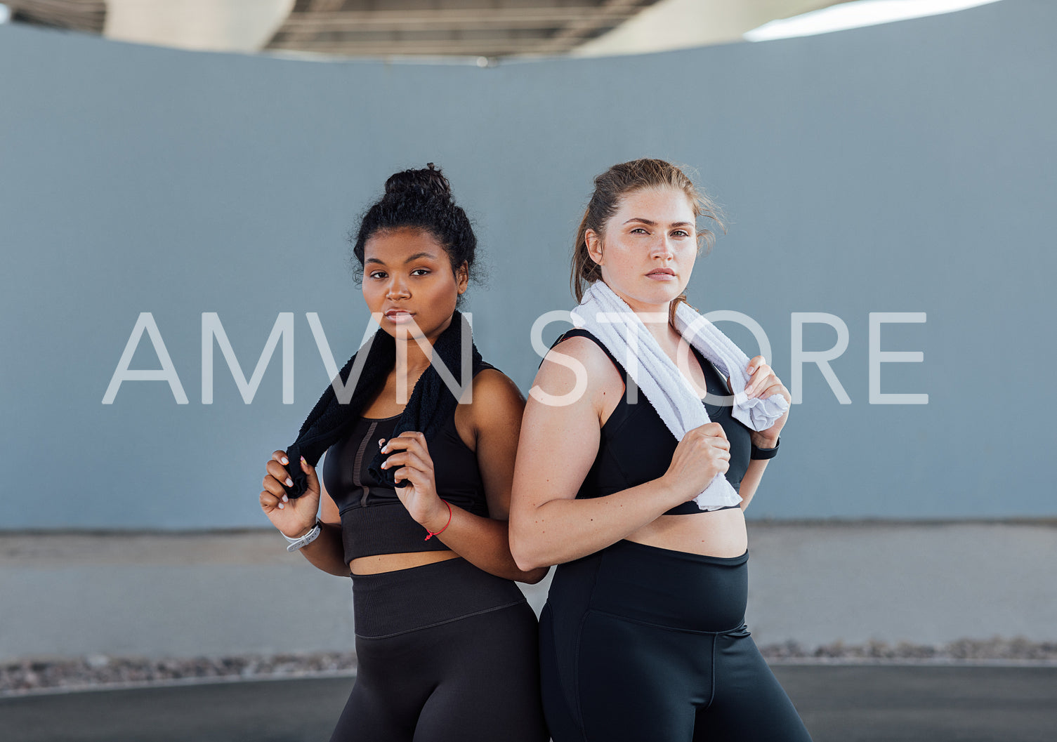 Two plus-size women in fitness wear standing back to back looking at camera. Two young females with towels posing after a workout.