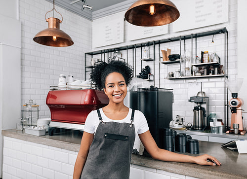 Young smiling woman with curly hair working as a barista. Female in an apron standing at a counter in a coffee shop and looking at camera.