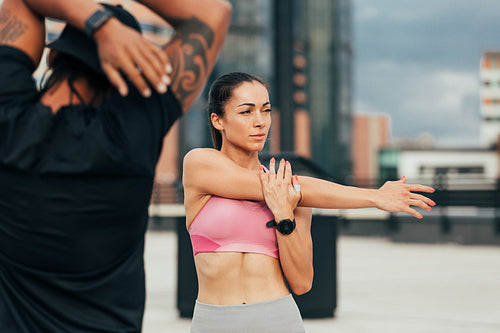 Two athletes are exercising on a rooftop. Young female stretching her arm before training.