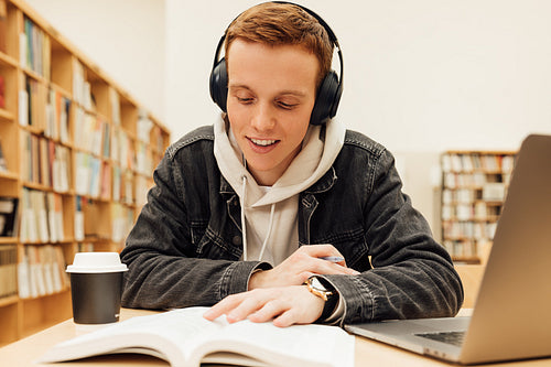 Smiling student reading a book wearing headphones while sitting in a library