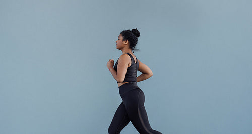 Side view of woman in black sportswear running at grey wall. Young female jogging outdoors.