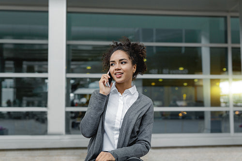 Front view of happy businesswoman sitting on bench and talking on mobile phone at airport terminal, looking away