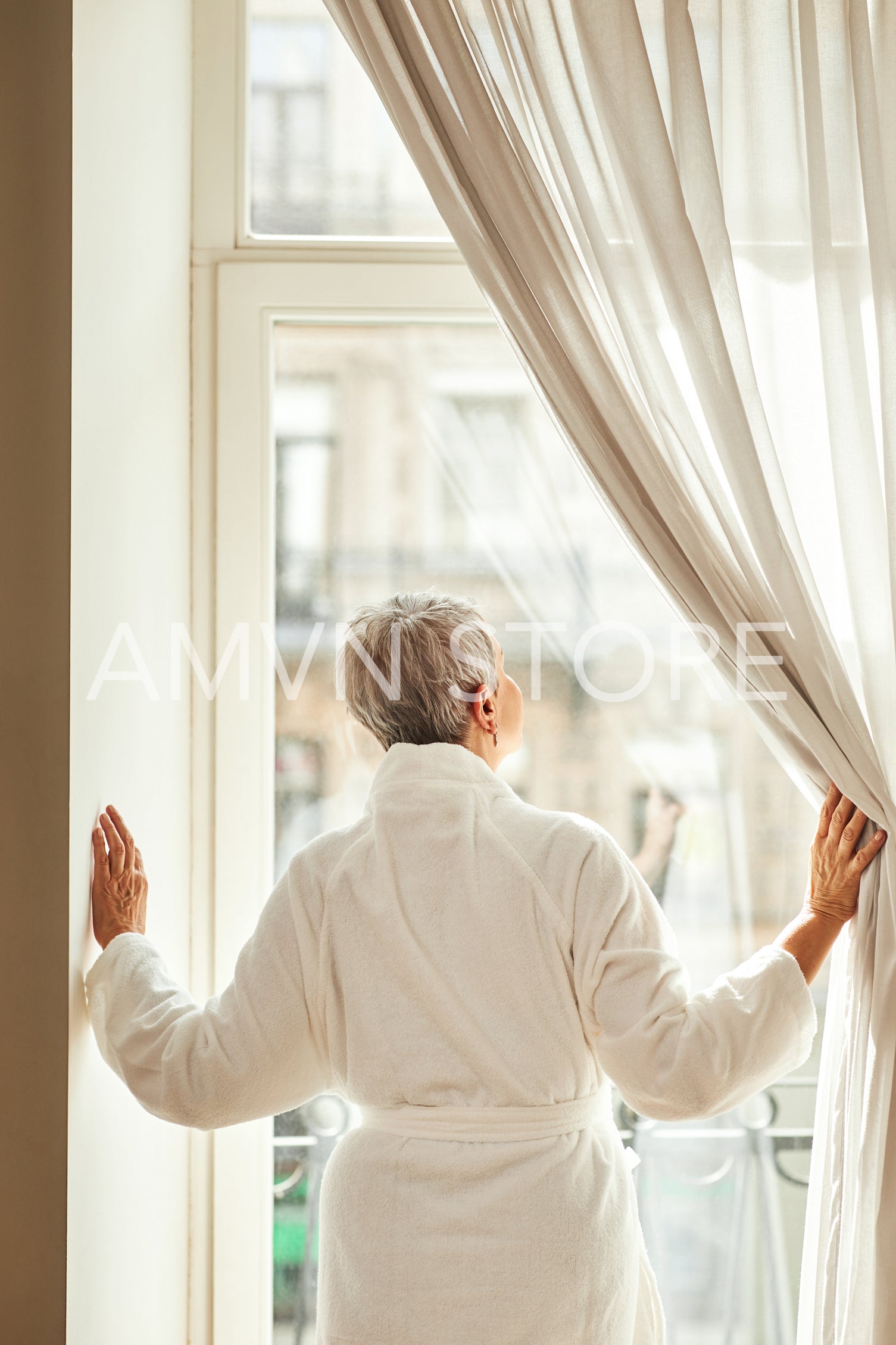 Rear view of a senior woman in bathrobe opening curtains in the morning	