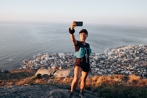 Woman hiker in sportswear taking selfie while standing on the top of the hill against cityscape