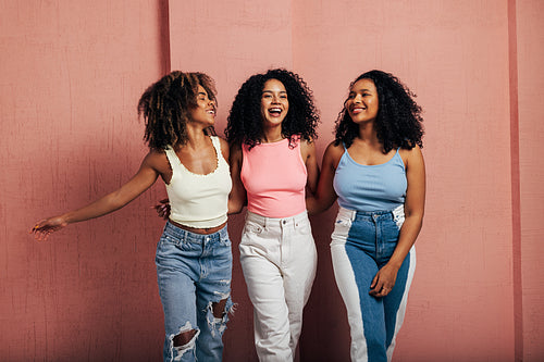 Three happy women with curly hair walk together. Young females having fun at a pink wall.