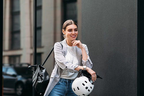 Smiling confident woman with an electrical scooter, safety helmet, and backpack standing at the building