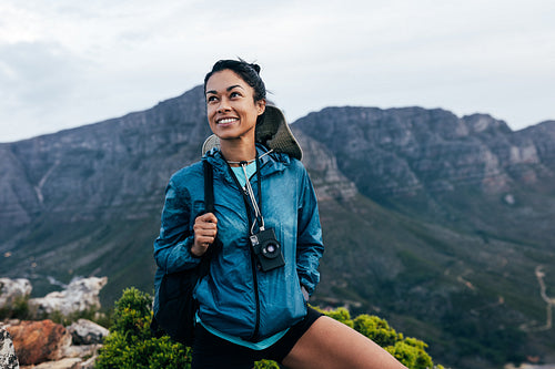 Portrait of a smiling woman relaxing during a mountain hike. Female with analog camera and backpack enjoying the view while standing on the top of the mountain.