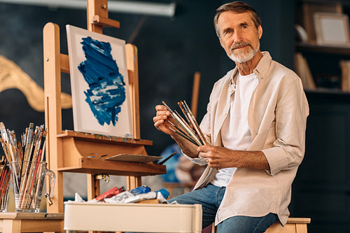 Male artist sitting with different paintbrushes in his hands and looking away