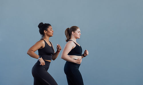 Side view of two females with similar body types running together at grey wall. Two women in black fitness attire running outdoors.