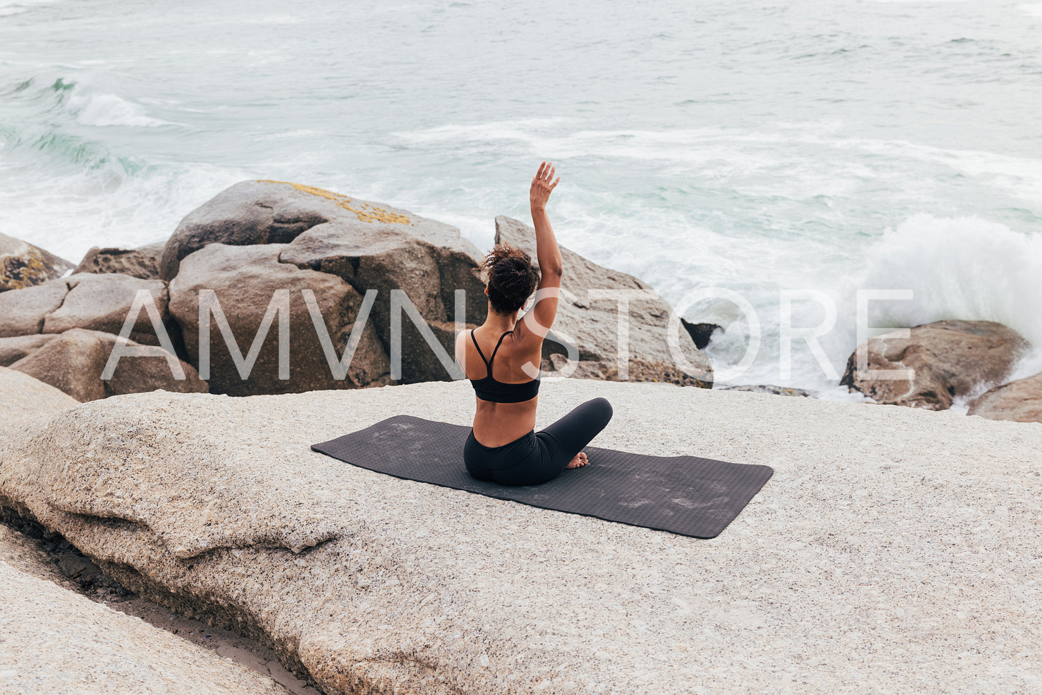 Rear view of fit female raising hand while changing yoga pose. Woman sitting on mat by shore and practicing yoga.