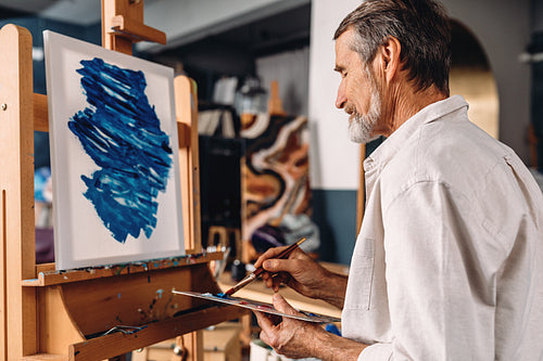 Concentrated bearded artist sitting in front of easel and mixing colors on palette with paintbrush