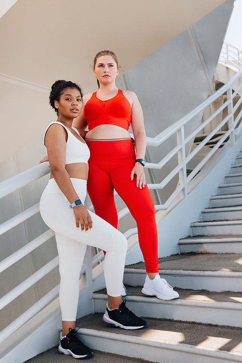 Full length of two plus-size females standing on stairs outdoors. Two young women in sportswear are looking away while taking a break during workout.