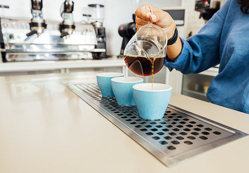 Hand of a female barista holding a glass teapot and pouring coffee into a small blue cup