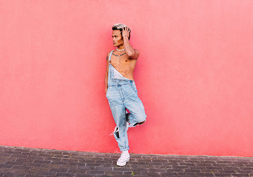 Stylish guy in casuals standing on one leg leaning on pink wall