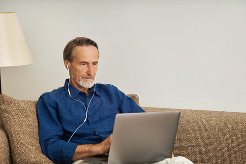 Senior man wearing earphones sitting on a couch with laptop