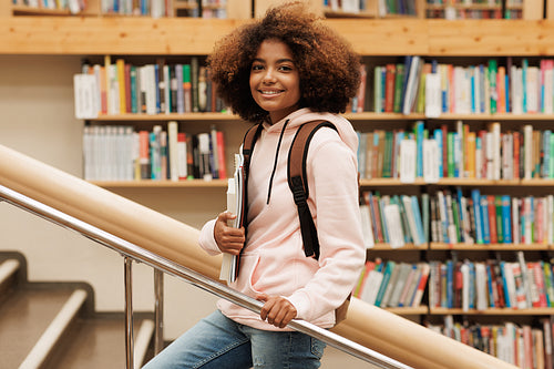 Beautiful girl with curly hair standing in library against bbokshelves and looking at camera