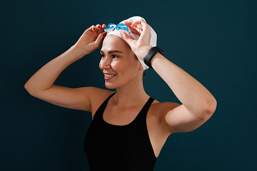 Portrait of a young female swimmer wearing goggles standing against a blue backdrop