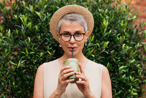 Stylish mature woman with grey hair drinking matcha latte outdoors