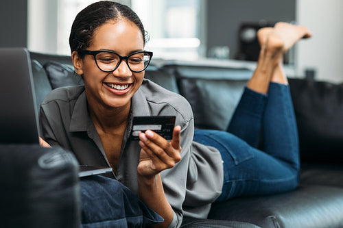 Happy young woman at home shopping online. Smiling female holding a credit card while lying on a sofa in a living room.