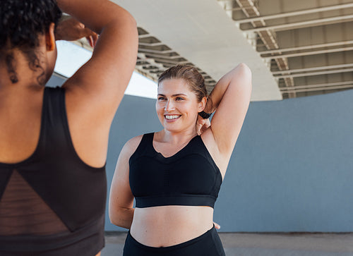 Smiling plus-size female doing warming up exercises outdoors looking at her fitness buddy