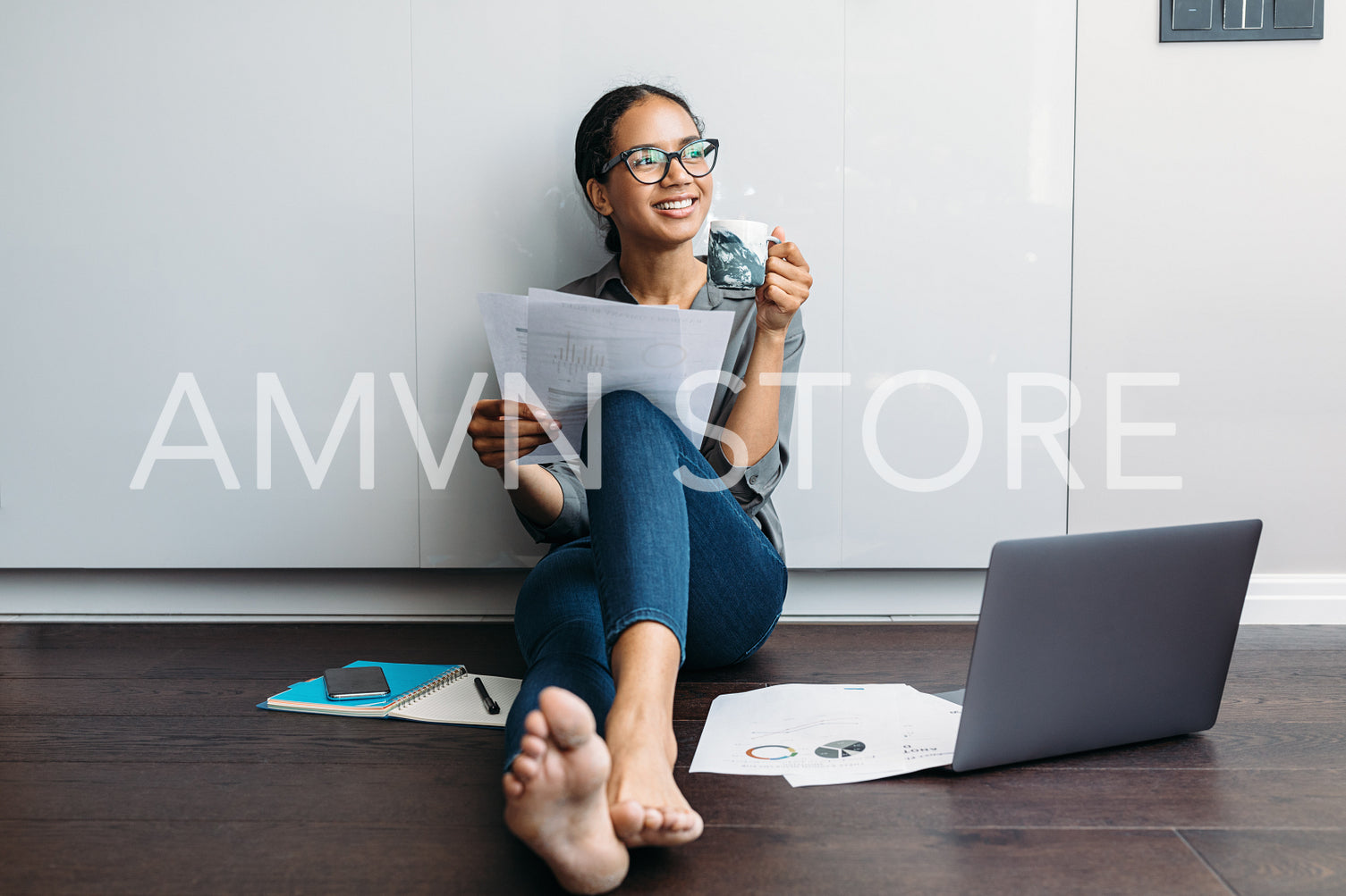 Smiling woman with a coffee mug holding documents. Young entrepreneur sitting on the kitchen floor looking away.	