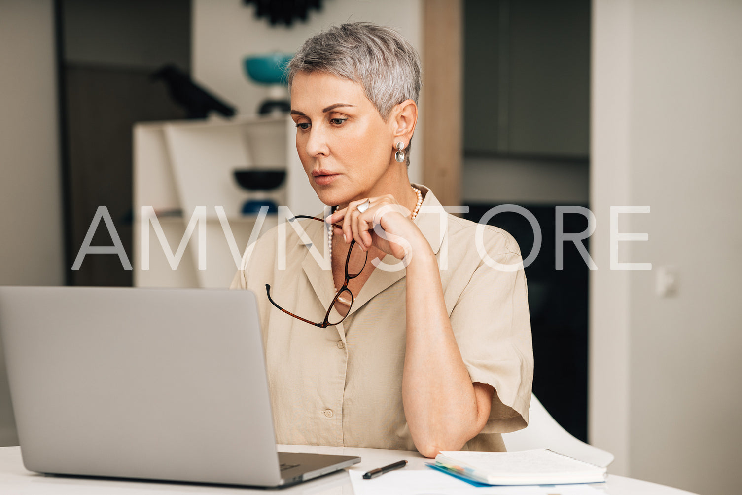 Mature woman in casuals sitting in the living room looking at laptop screen and holding spectacles