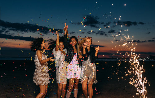 Happy friends celebrating outdoors at sunset. Four stylish women throwing confetti outdoors near fireworks.