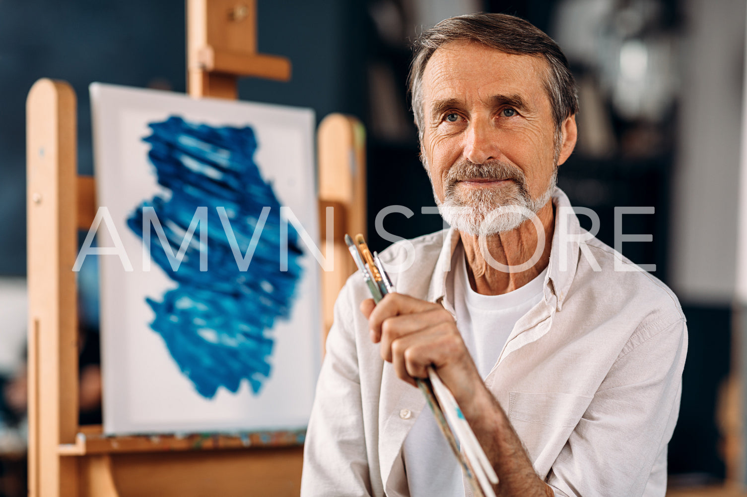 Portrait of bearded painter sitting against his picture and looking away	