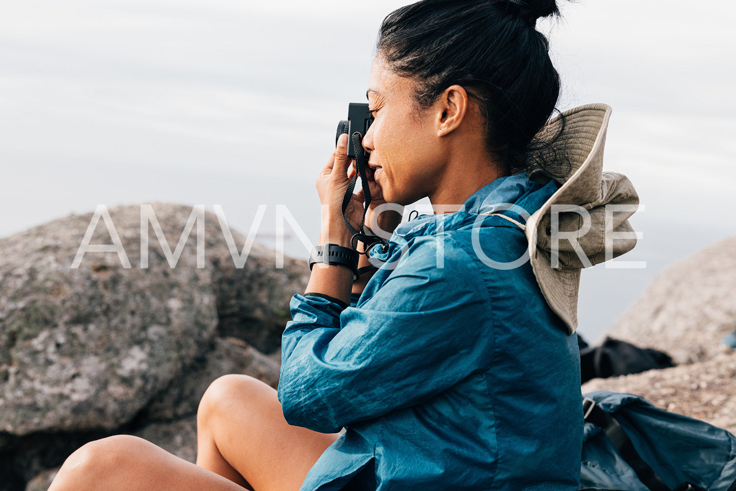 Side view of a woman taking photographs on her film camera while sitting on a rock during a mountain hike